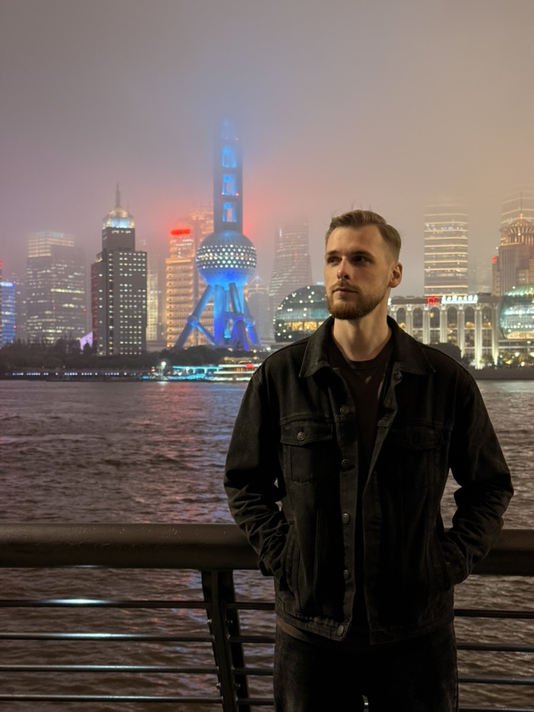 Portrait of Vitalii Davydov standing by a river embankment at night, with an illuminated city skyline in the background